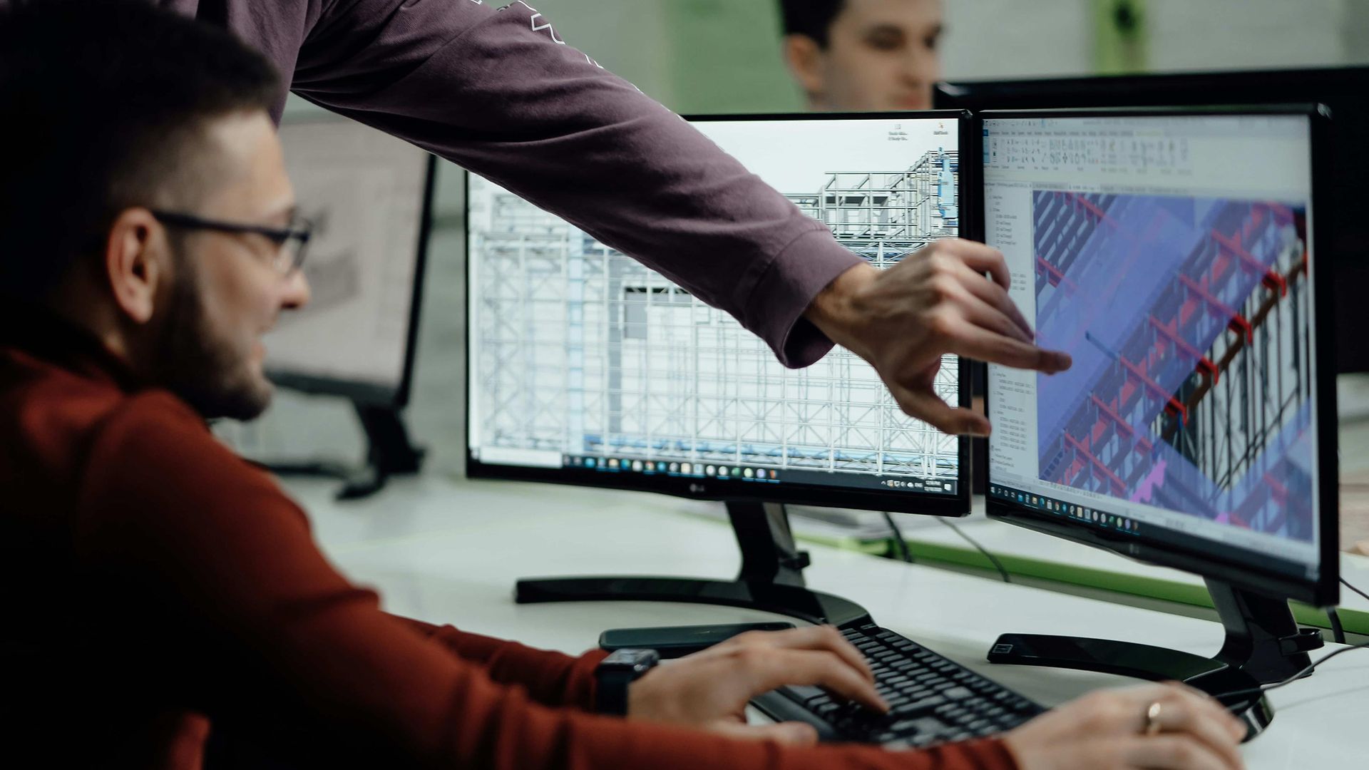 Ein Mann sitzt am Schreibtisch vor zwei Bildschirmen. Eine weitere Person zeigt ihm etwas am Bildschirm per Hand
