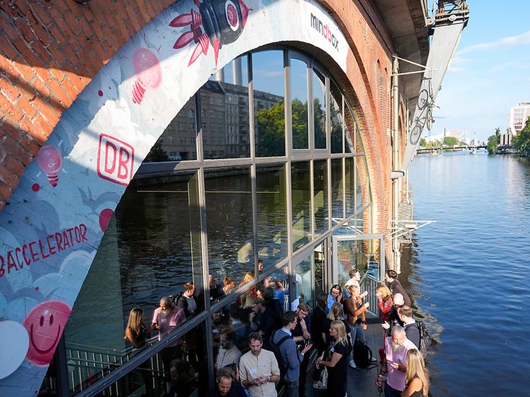 Several people are standing outside the DB Mindbox by the water.