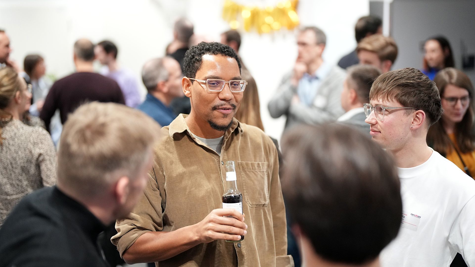 A man is holding a drink and looking toward the camera, while several people are standing around him.