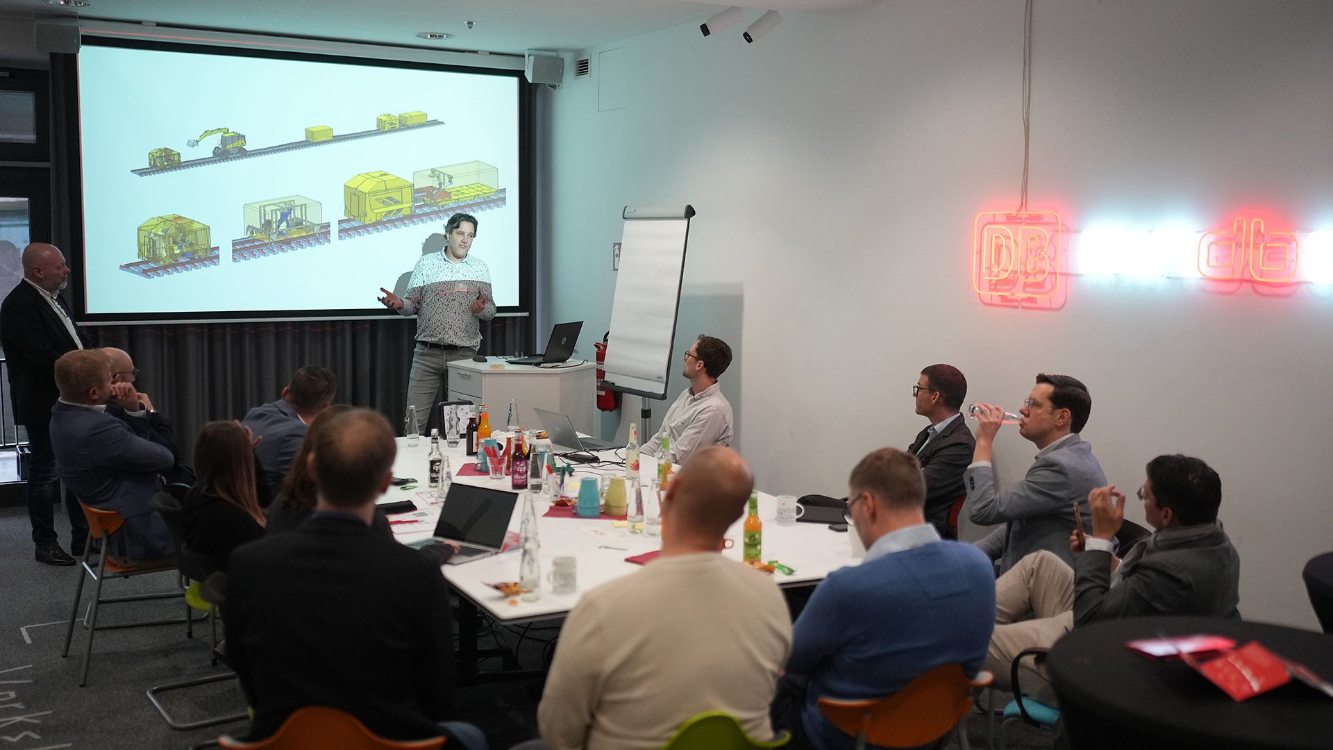 Several people are seated in a meeting room, listening to a presentation.